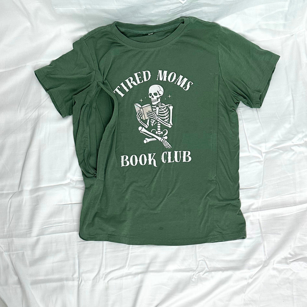 An olive green short-sleeve t-shirt with a white graphic print that reads 'TIRED MOMS BOOK CLUB' around a skeleton holding a book, laid flat on a wrinkled white sheet.