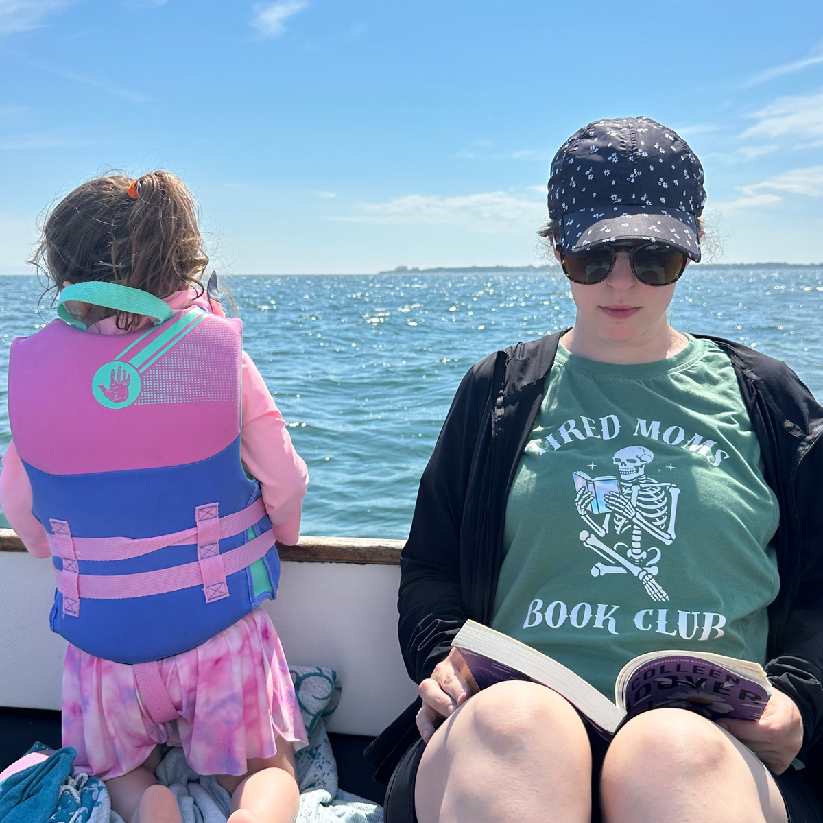 A woman and a child sitting on a boat outdoors by the water. The child wears a pink and blue life vest, and the woman wears a green graphic T-shirt that reads 'TIRED MOMS BOOK CLUB' with a skeleton illustration.