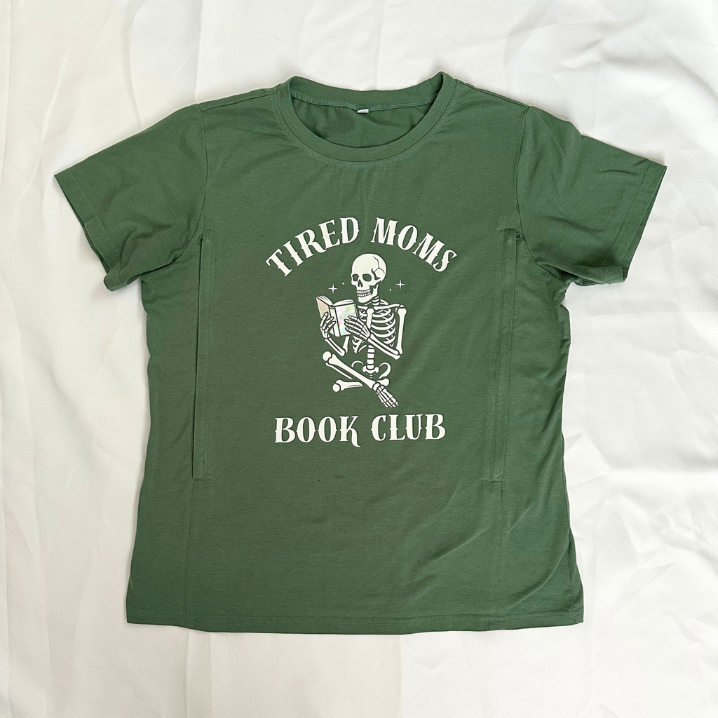 An olive green short-sleeve t-shirt laid flat on a white fabric background, featuring a white graphic of a skeleton holding a book with the text 'TIRED MOMS BOOK CLUB'.