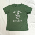An olive green short-sleeve t-shirt laid flat on a white fabric background, featuring a white graphic of a skeleton holding a book with the text 'TIRED MOMS BOOK CLUB'.
