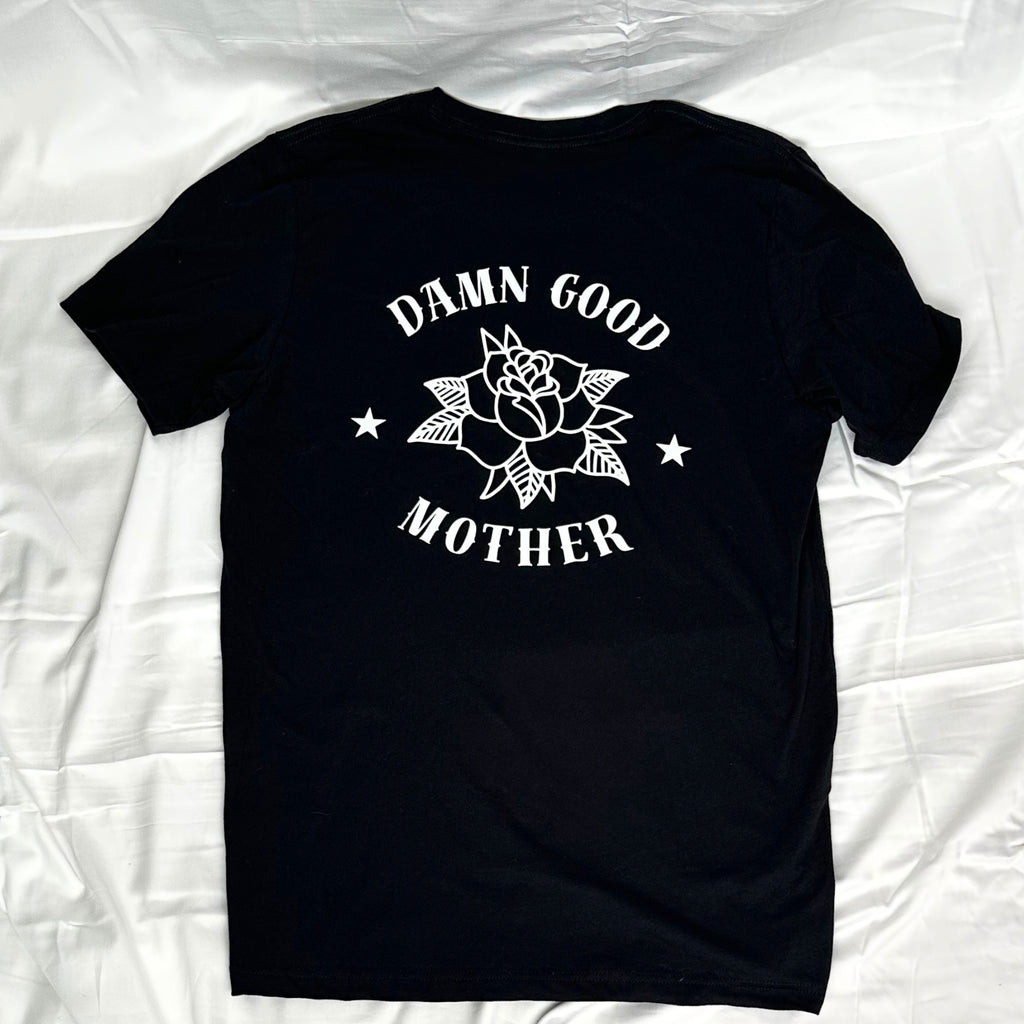 A black short-sleeve t-shirt laid flat, featuring a white graphic print on the back with a rose, stars, and the text 'DAMN GOOD MOTHER'.