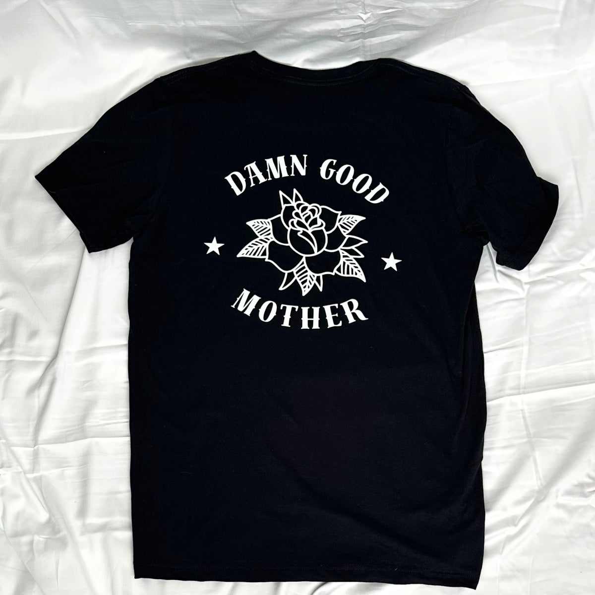 A black short-sleeve t-shirt laid flat, featuring a white graphic print on the back with a rose, stars, and the text 'DAMN GOOD MOTHER'.