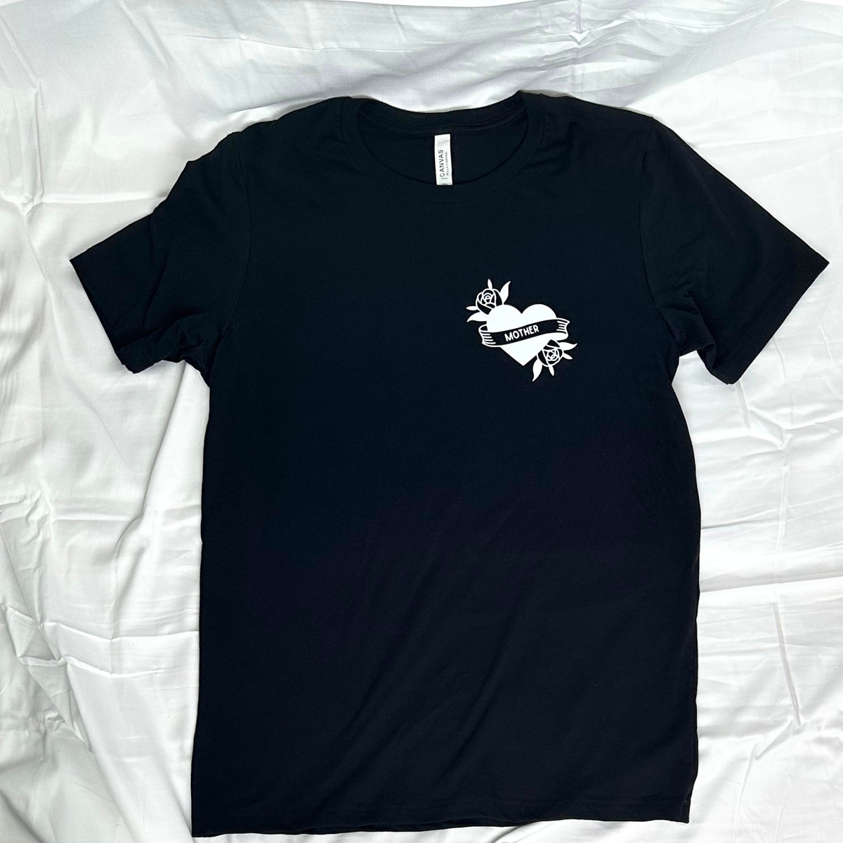 A black short-sleeve t-shirt with a small white graphic on the left chest featuring a heart, ribbon, and flowers, laid flat