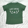A green short-sleeve crewneck t-shirt laid flat on a white wrinkled fabric background. The shirt features a white graphic print on the chest that reads 'EAT LOCAL' above a decorative floral and starburst pattern.