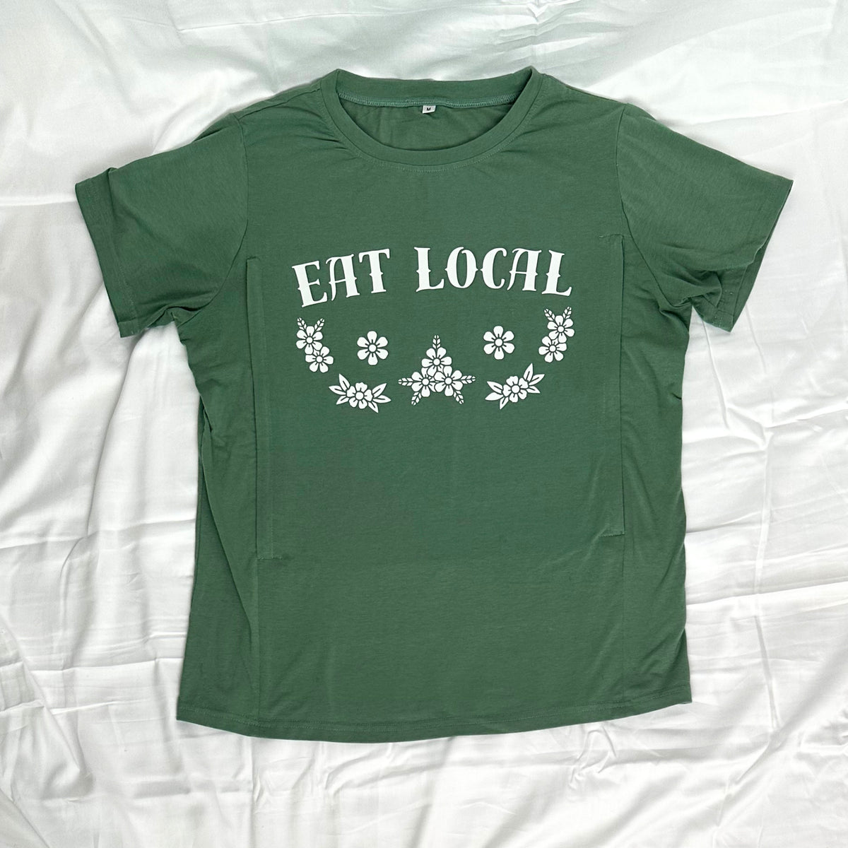 A green short-sleeve crewneck t-shirt laid flat on a white wrinkled fabric background. The shirt features a white graphic print on the chest that reads 'EAT LOCAL' above a decorative floral and starburst pattern.