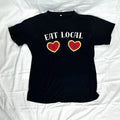 A black short-sleeve t-shirt laid flat. The shirt features the words 'EAT LOCAL' in white across the chest, with two red hearts outlined in yellow below the text.
