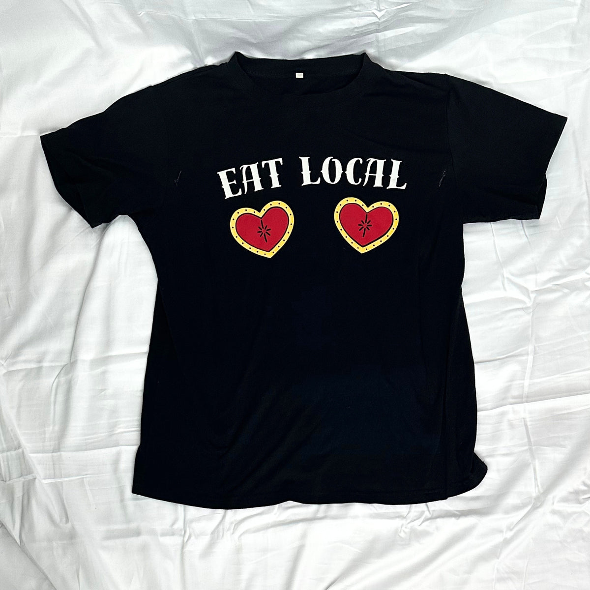 A black short-sleeve t-shirt laid flat. The shirt features the words 'EAT LOCAL' in white across the chest, with two red hearts outlined in yellow below the text.