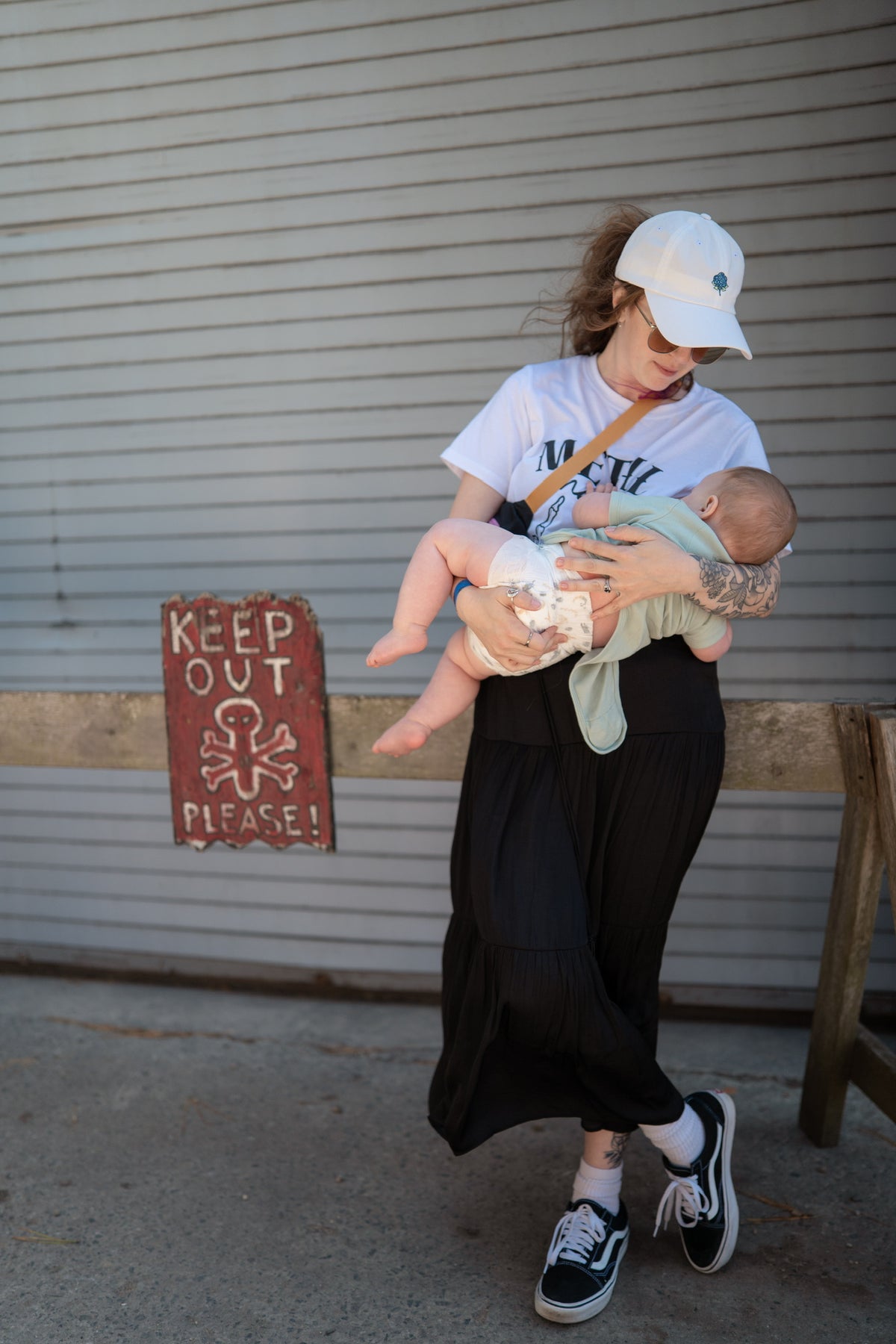 One of our founders, Nic, a lady with alternative/witchy style nursing a baby in front of a metal shutter with a 'Keep Out' sign, wearing Metal Mama nursing tee.