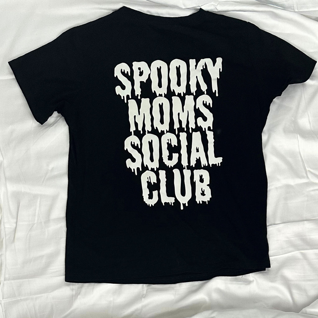 A black short-sleeve t-shirt laid flat on white fabric, featuring the phrase 'SPOOKY MOMS SOCIAL CLUB' printed in a large, dripping white font on the back.