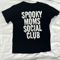 A black short-sleeve t-shirt laid flat on white fabric, featuring the phrase 'SPOOKY MOMS SOCIAL CLUB' printed in a large, dripping white font on the back.