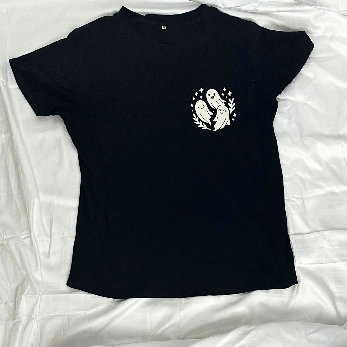 A black short-sleeve t-shirt laid flat, featuring a small white graphic on the left chest of three ghosts inside a circular wreath with stars.